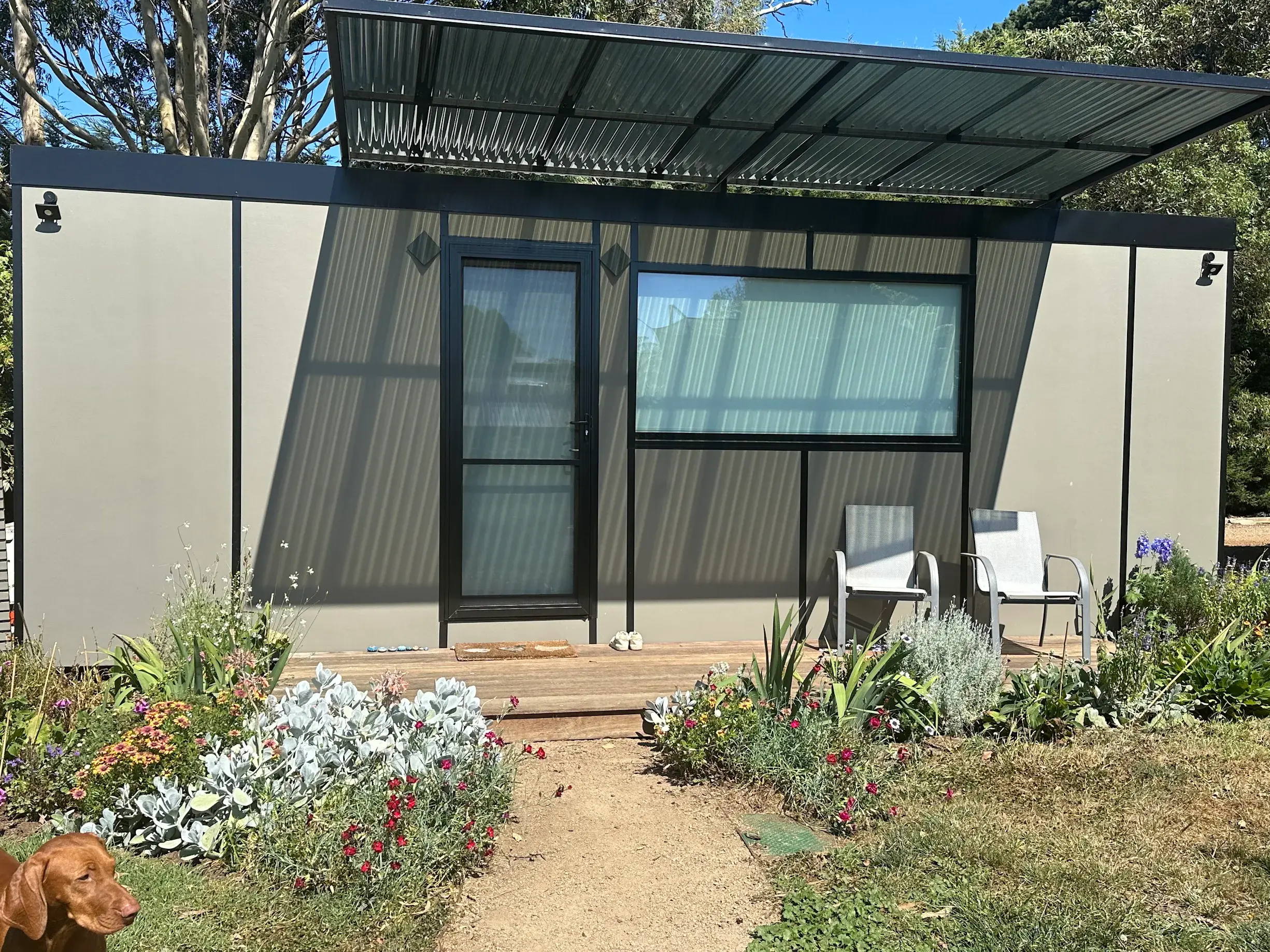 Anne Altmann’s studio MicroHome behind a flowering garden bed, with a black-framed door and picture window under a cantilevered awning.