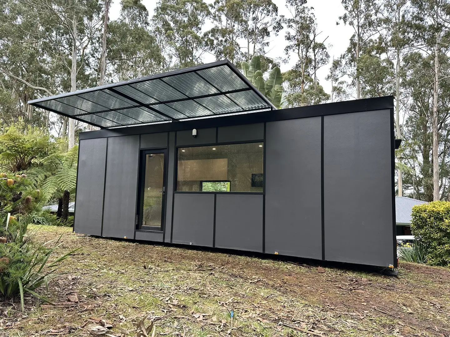 A charcoal MicroHome with a raised solar awning, sited on a sloping bush block among tree ferns and gums.