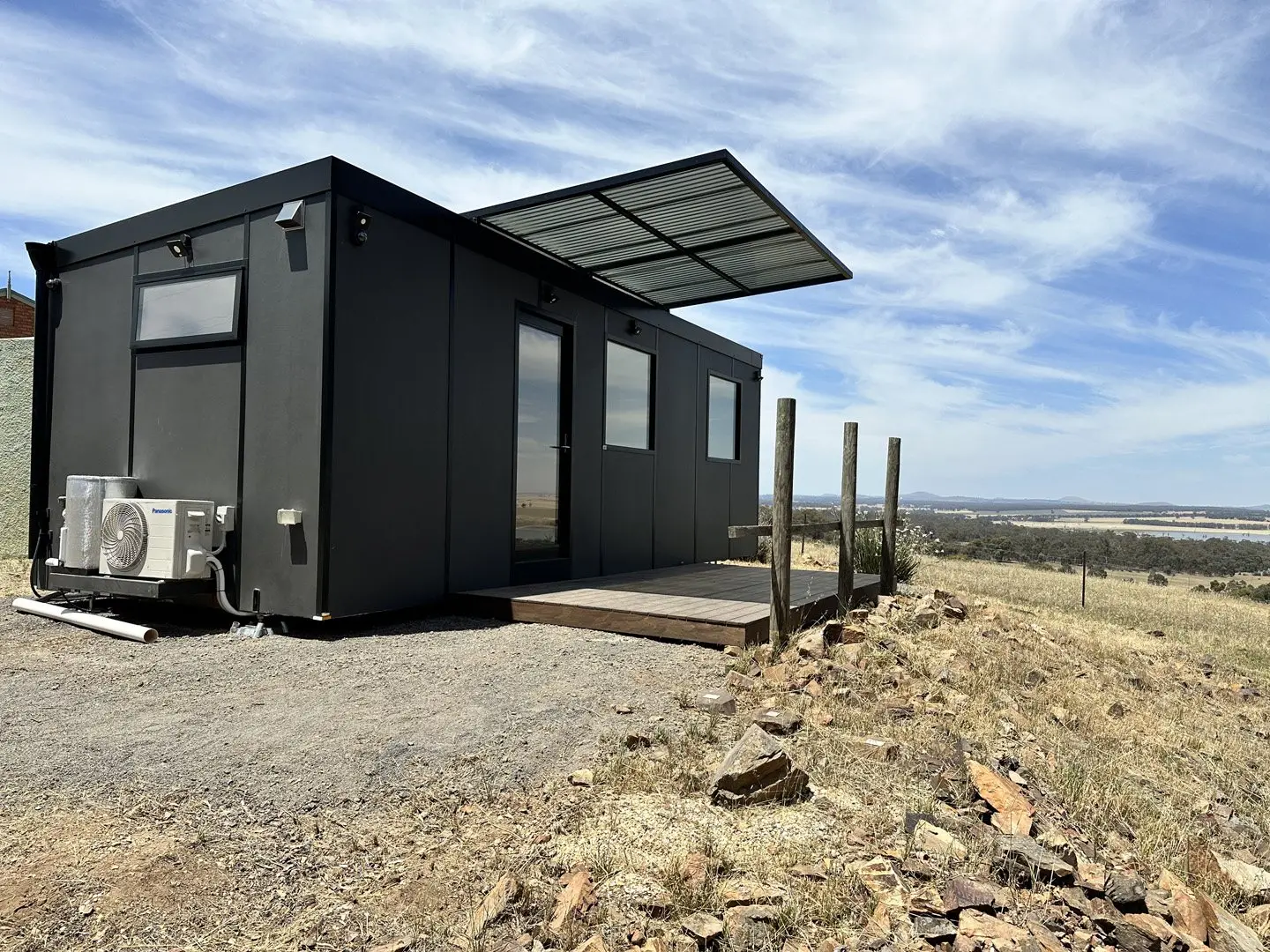 A charcoal MicroHome with a timber entry deck, sited on a hillside block with open country behind it.