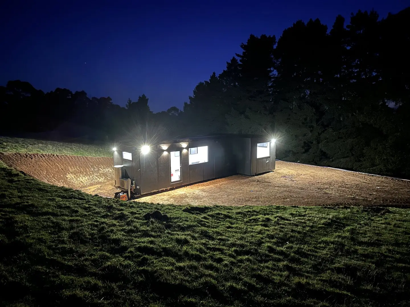A charcoal MicroHome lit from within at night on a cut pad in a paddock, tall trees behind it.