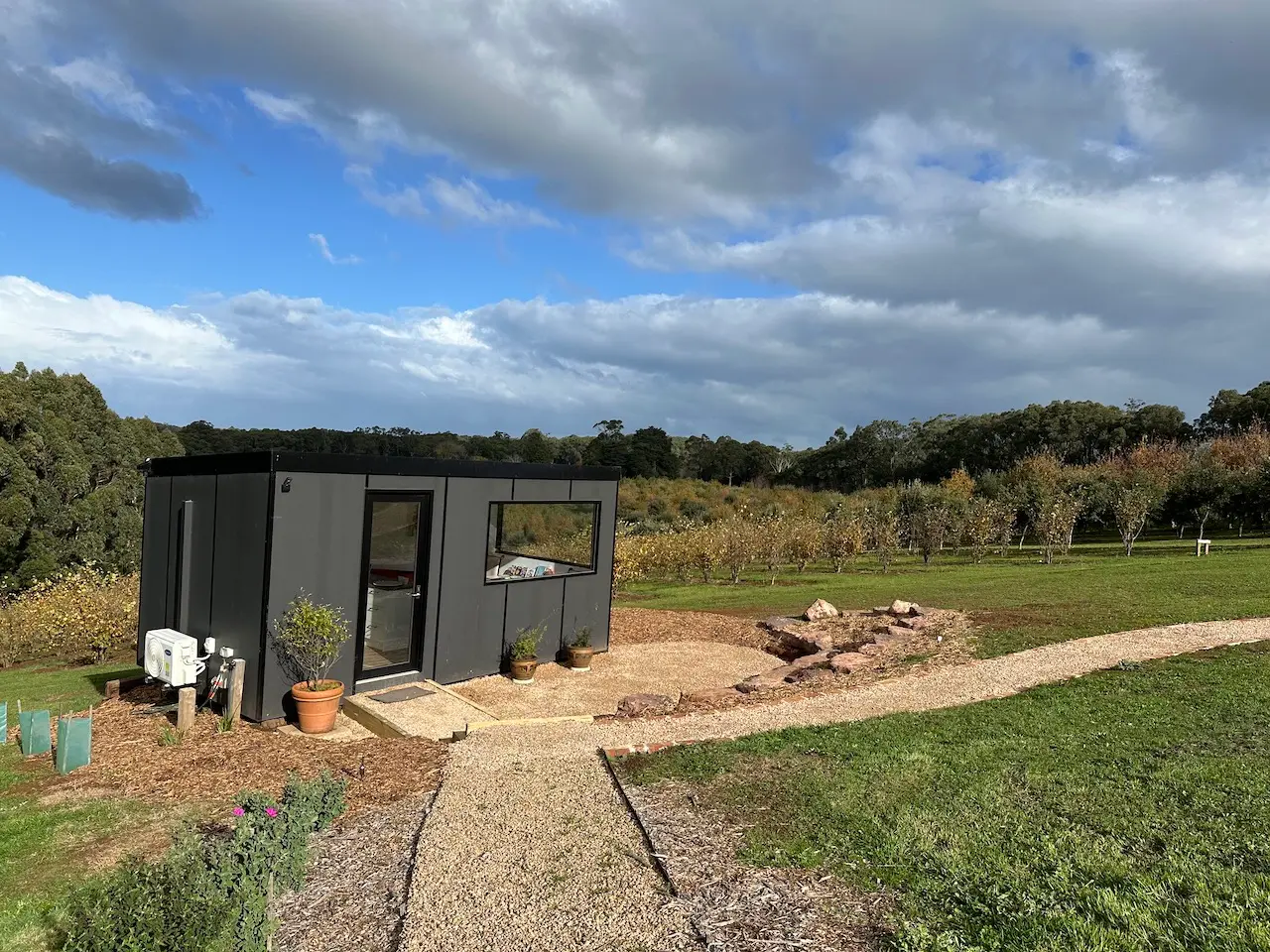 A charcoal-clad MicroHome sited on a grassy rise above an orchard, with a gravel path leading to its door under a broken blue sky.