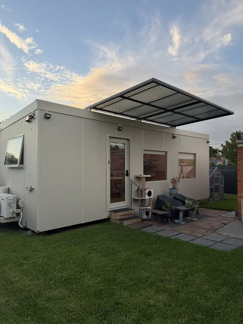 A cream MicroHome with a raised solar awning, sited on the lawn of a suburban backyard with a paved sitting area outside its door.