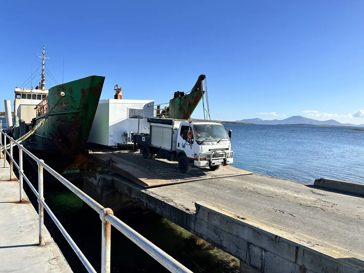 A MicroHome and its crane truck being delivered by barge at a jetty on the water.