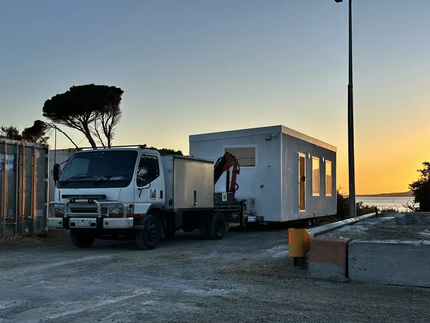 A MicroHome on the back of a MicroHomes crane truck at a coastal landing at sunset.