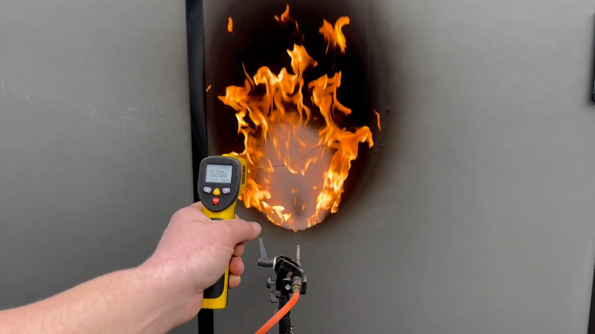 A flame held against MicroHome wall cladding during a fire test, with a temperature probe alongside.