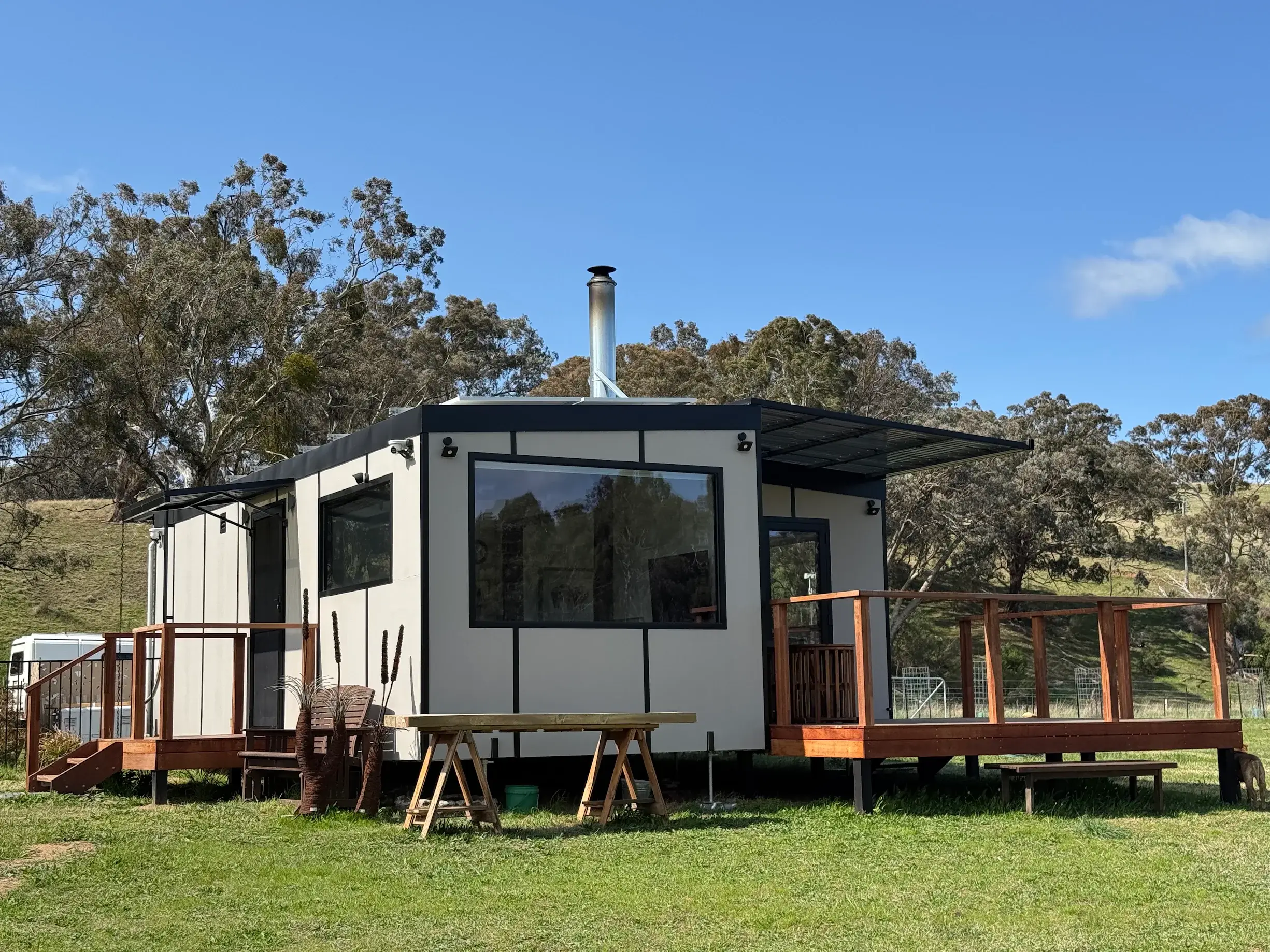 Heather James’s white MicroHome on a rural block, with a flue and a timber deck, hills behind.