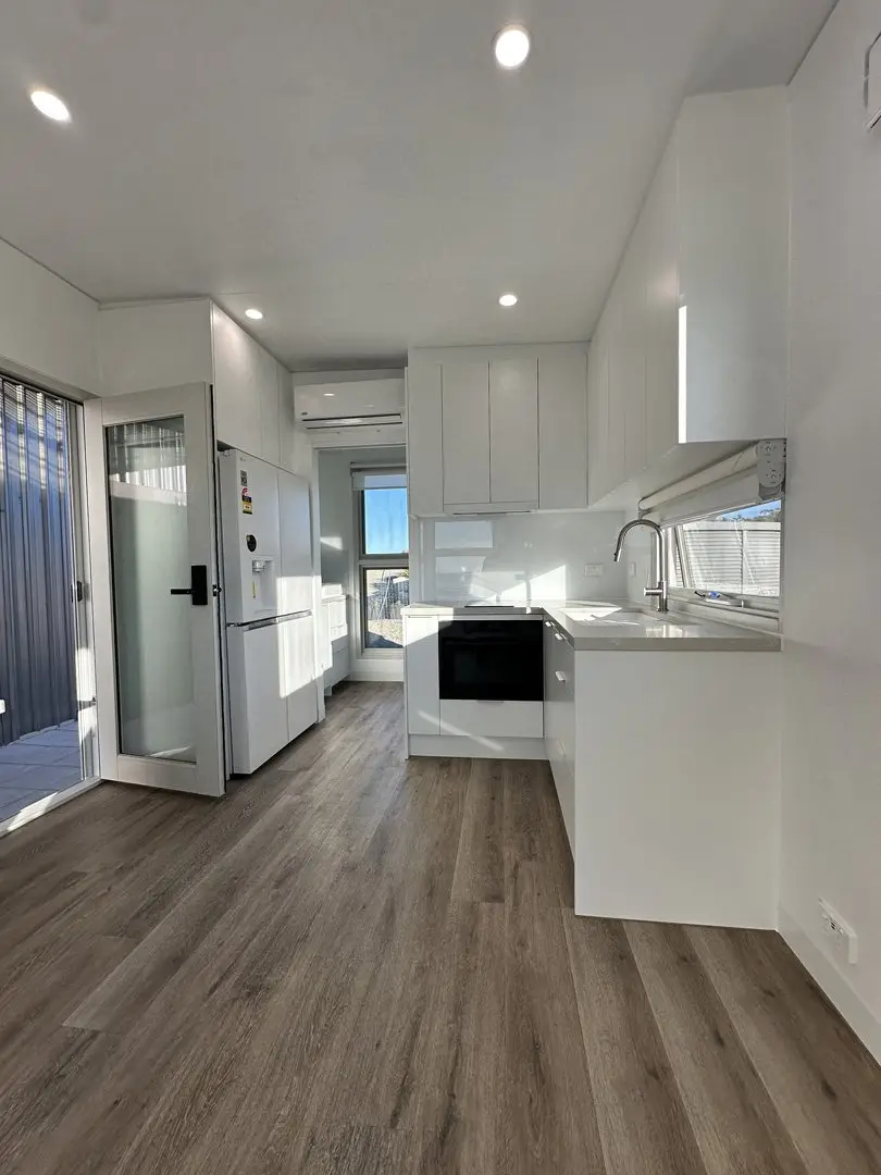 Looking down the length of a white-finished MicroHome interior: a kitchen with a fridge and oven, a glazed door and a timber-look floor.