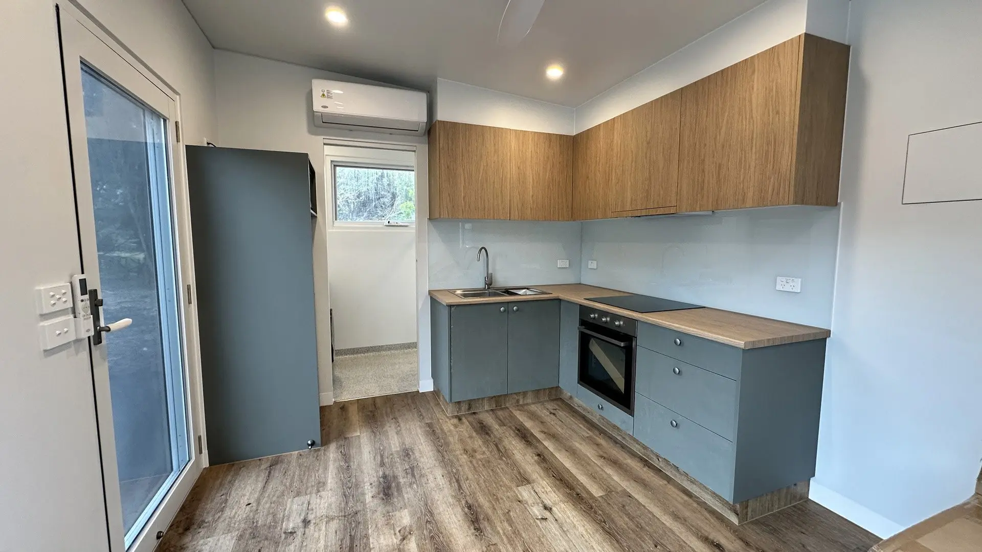 A MicroHome kitchen with timber-look overhead cabinets, grey-blue base cabinets and an oven, beside a glazed door with a reverse-cycle split system above it.