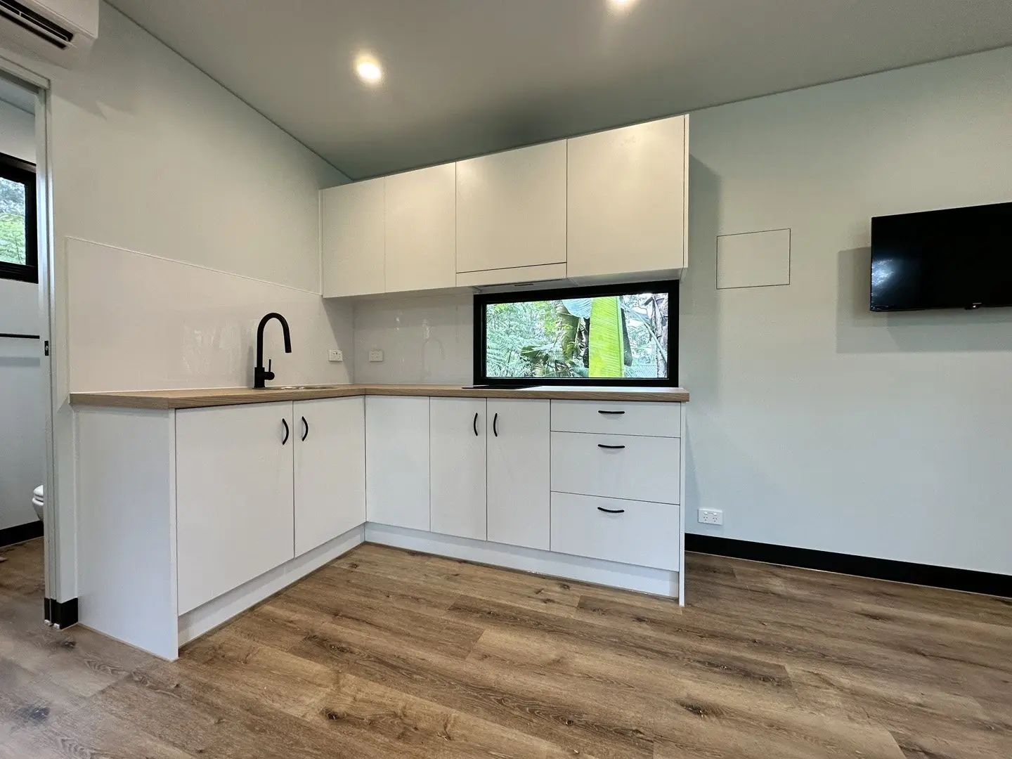 A white corner kitchen in a MicroHome with a timber-look bench, a black tap, a window over the sink and a wall-mounted TV.