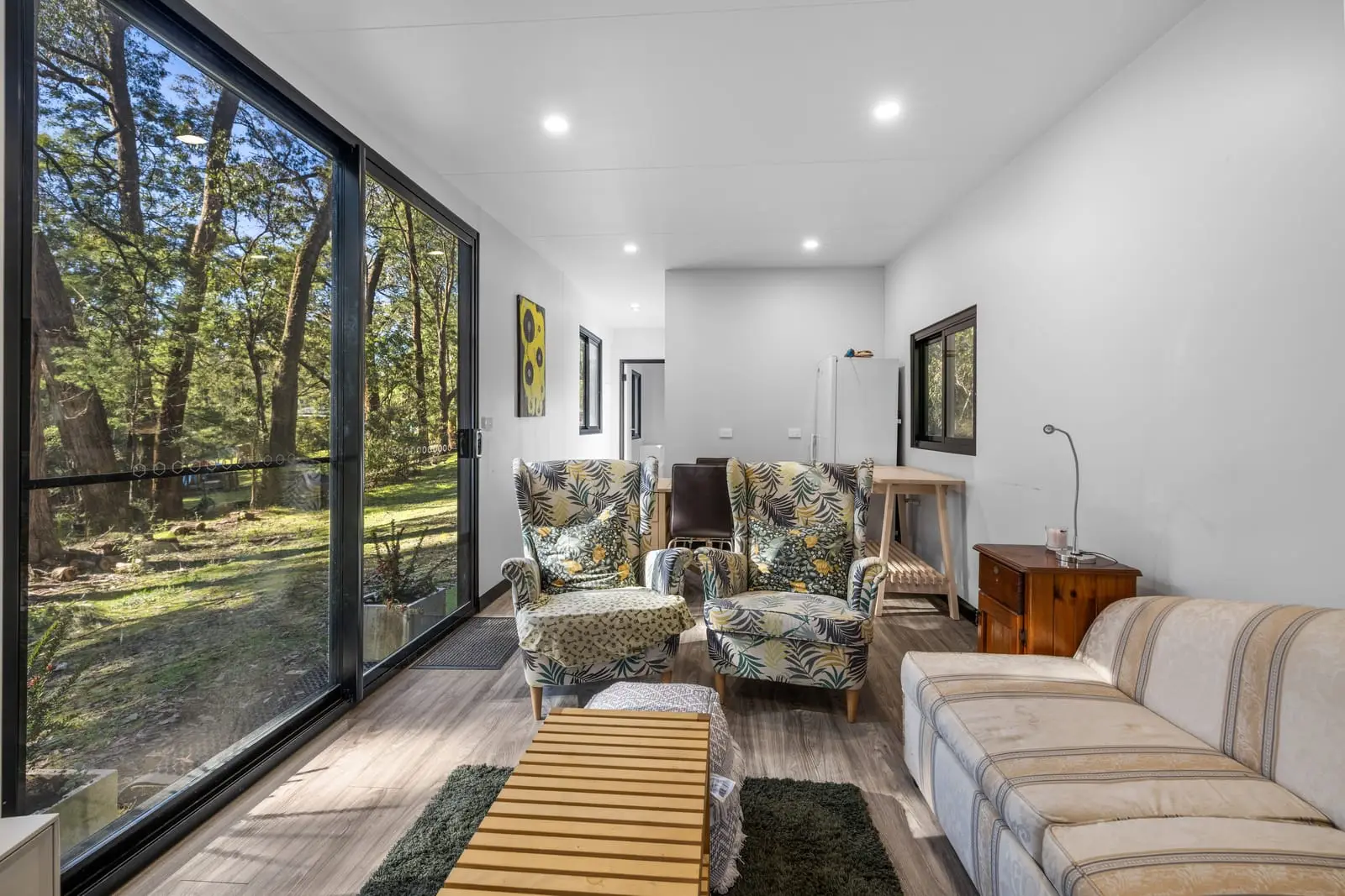 The living room of a finished MicroHome, with a full-height window wall looking into bushland, armchairs and a sofa.