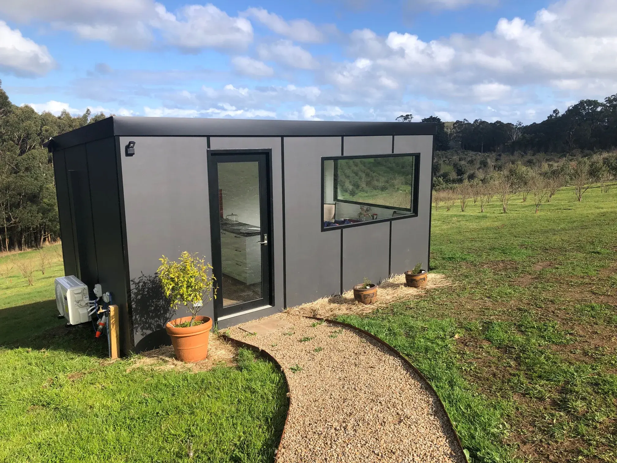 Mike McAuley’s charcoal MicroHome set level on a rural block, a gravel path to the door and an orchard beyond.