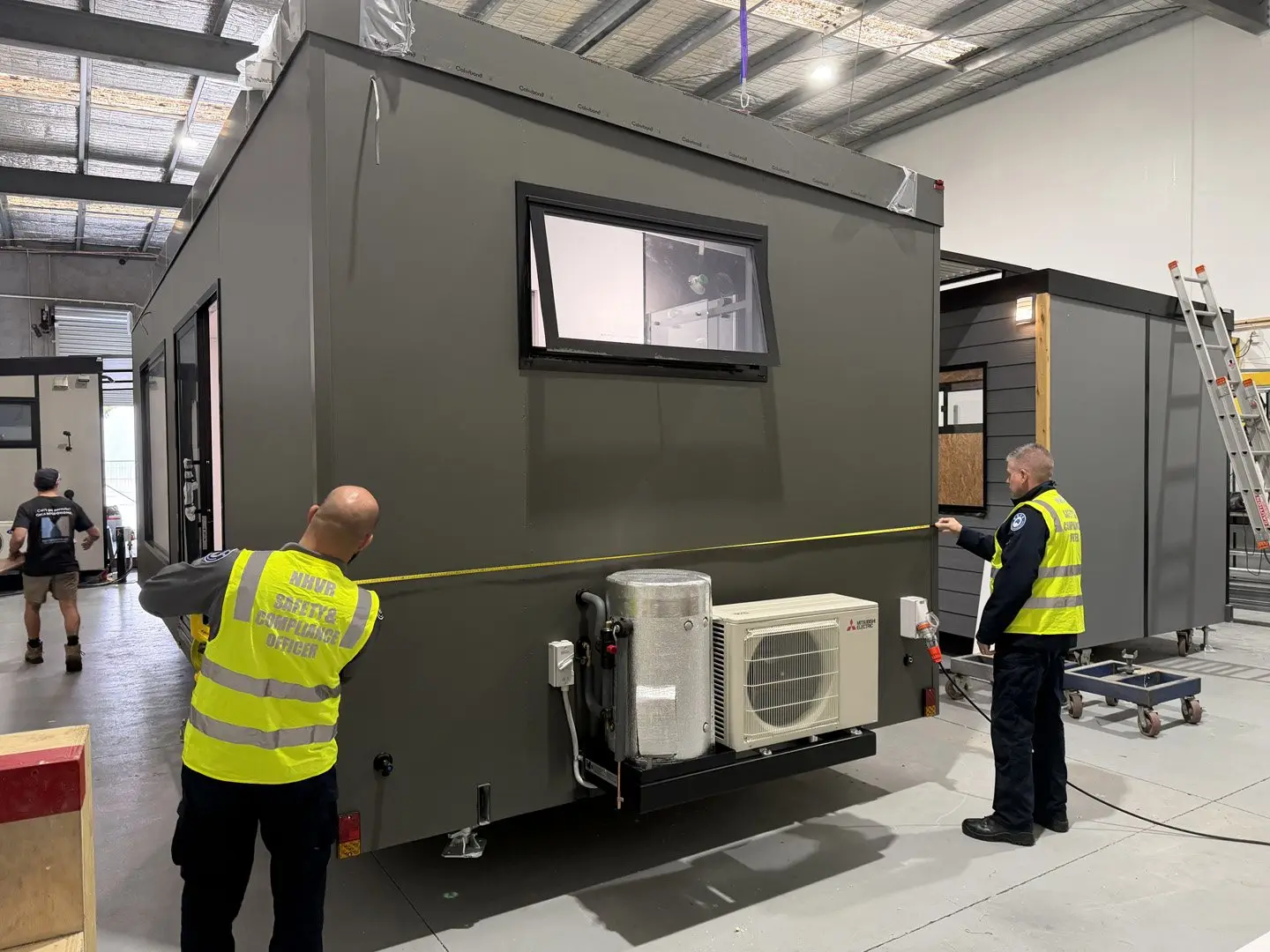 An officer from the National Heavy Vehicle Regulator in a hi-vis vest measuring a MicroHome with a tape at the MicroHomes factory.