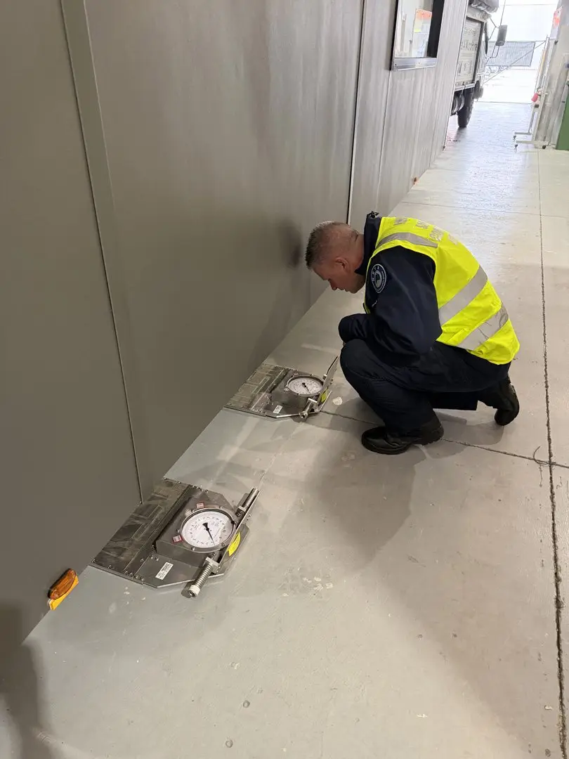 An officer in a hi-vis vest crouched beside the wall of a MicroHome, reading dial gauges placed on the factory floor.