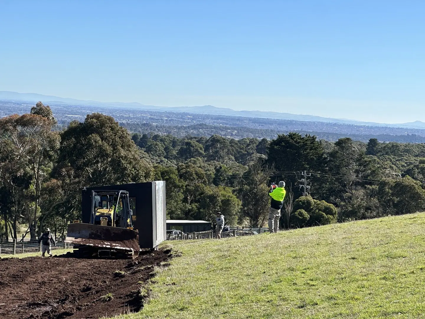 A MicroHome being positioned by a dozer on a cut pad on a hillside, with the valley below.