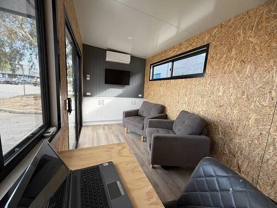 Inside a Backyard Pod fitted out as a quiet home office — an OSB feature wall, a wall-mounted screen, soft seating and a wide window onto the yard.