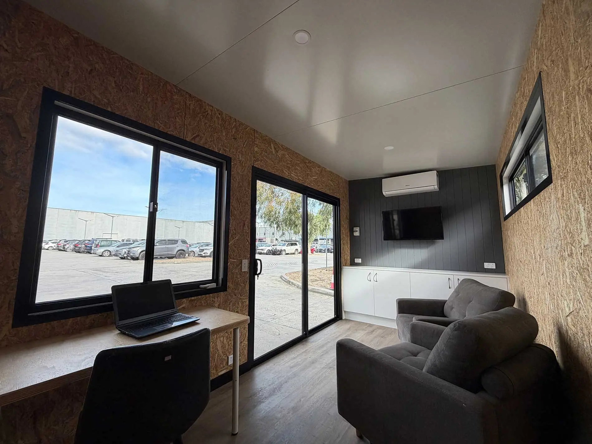 The full length of a Backyard Pod interior — soft armchairs along an OSB feature wall, a wall-mounted screen and a split-system head, opening to the yard through full-height sliding doors.