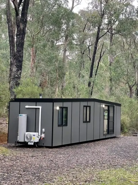A MicroHome sited among trees, with its external hot-water unit and services along the side.