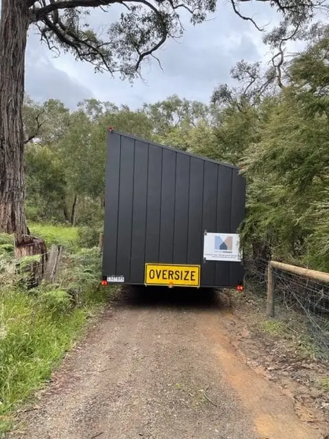 A MicroHome on its chassis being towed up a narrow dirt track behind a vehicle, with an OVERSIZE sign on the rear.